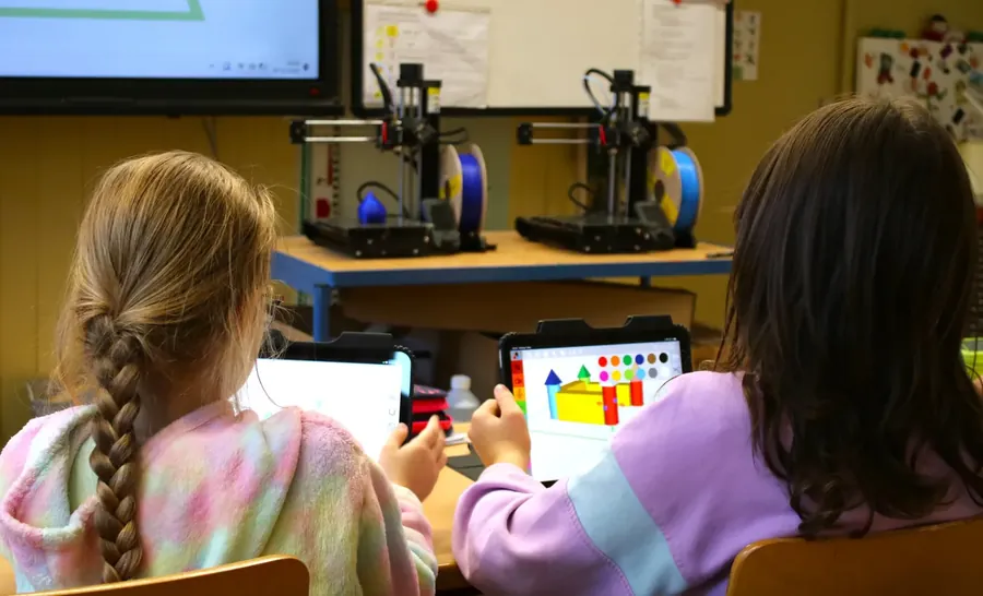 Zwei Schülerinnen konstruieren am Tablet eine bunte Burg, im Hintergrund stehen zwei 3D-Drucker