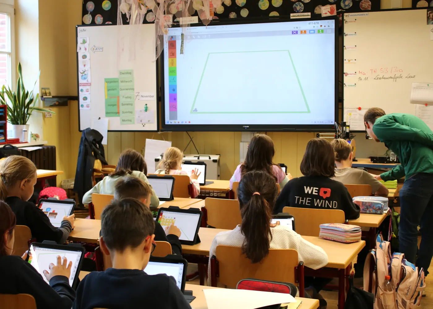 Eine Grundschulklasse arbeitet an Tablets mit TinkerSchool, vorne zeigt das digitale Board den Baukasten, die Lehrkraft hilft an einem Tisch